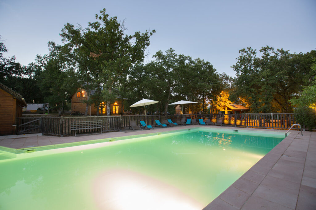 Piscine illuminée au cœur du domaine, entourée de chalets en bois et d'arbres centenaires pour une pause détente inoubliable