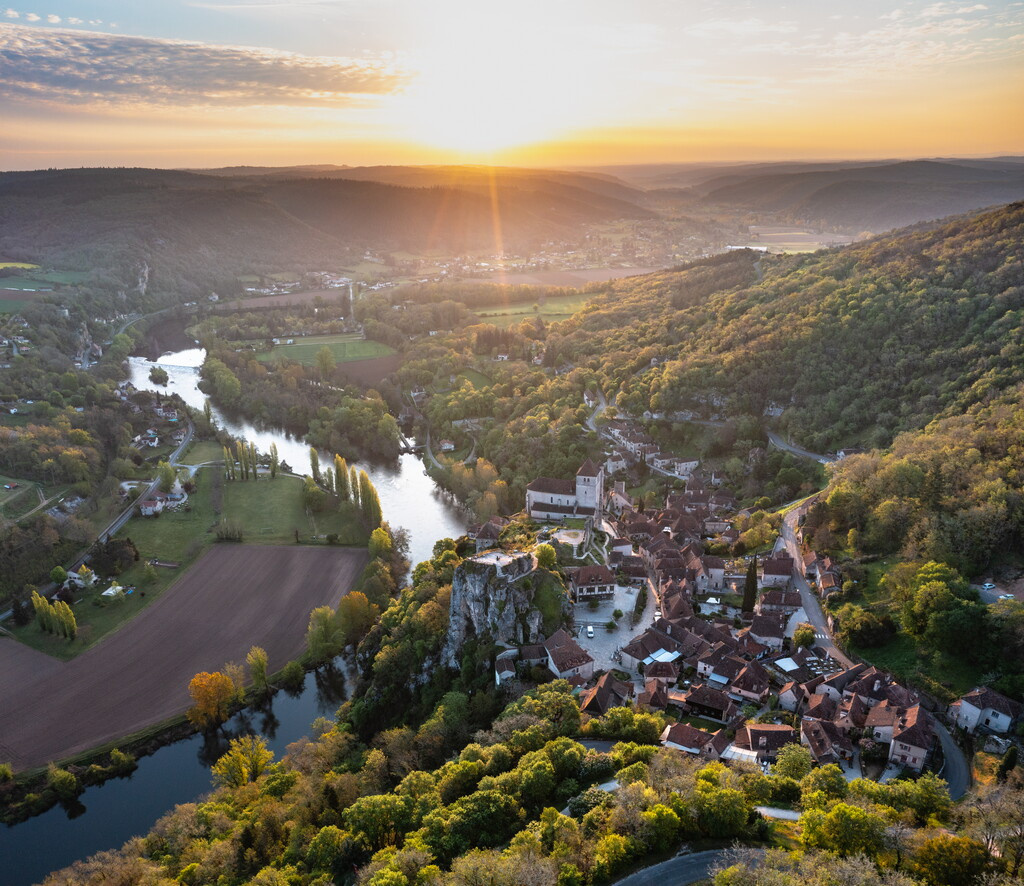 Vue majestueuse sur la vallée du Lot au coucher de soleil, avec Saint-Cirq-Lapopie perché sur sa falaise, forêts verdoyantes et rivière sinueuse